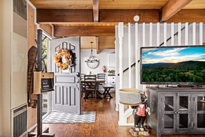 A cozy living room with a wooden ceiling, a TV on a cabinet, a dining area with a chandelier, and a decorative wreath on the door.