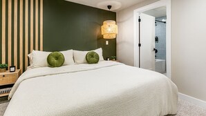 Un dormitorio con cama, mesita de noche, lámpara de pared y puerta que da a un baño.