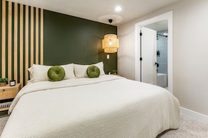 Un dormitorio con cama, mesita de noche, lámpara de pared y puerta que da a un baño.