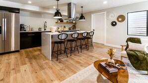 Una cocina moderna con una isla central, taburetes de bar y una zona de comedor con una silla verde y una mesa de centro.