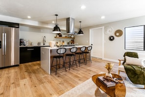 Una cocina moderna con una isla central, taburetes de bar y una zona de comedor con una silla verde y una mesa de centro.