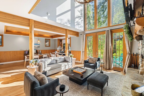 A spacious living area with a high ceiling, wooden beams, large windows, and a stone fireplace.