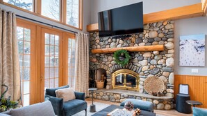 A cozy living room with a stone fireplace, a flat-screen TV mounted above it, and a large window with curtains. - Palisades Cabin Retreat | Hot Tub•A/C•EV•Views (Alpine Meadows)