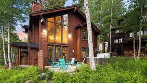A wooden cabin with large windows, a deck with chairs, and a forested backdrop. - Palisades Cabin Retreat | Hot Tub•A/C•EV•Views (Alpine Meadows)