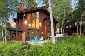A wooden cabin with large windows, a deck with chairs, and a forested backdrop.