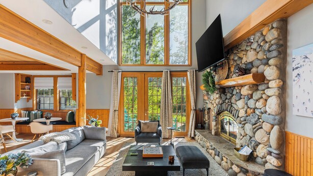 A spacious living room with a stone fireplace, wooden beams, and large windows. - Palisades Cabin Retreat | Hot Tub•A/C•EV•Views (Alpine Meadows)