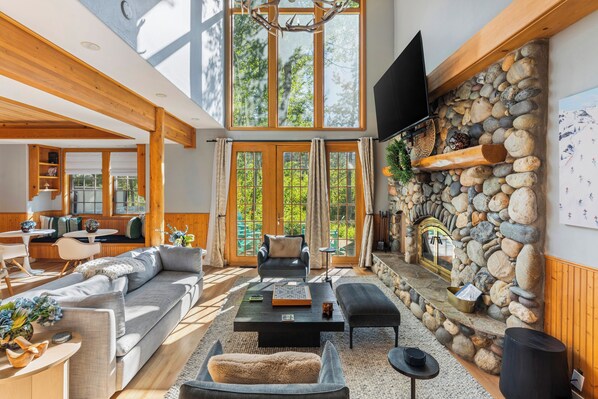 A spacious living room with a stone fireplace, wooden beams, and large windows.
