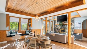 A wooden interior with a dining area, stone fireplace, and large windows. - Palisades Cabin Retreat | Hot Tub•A/C•EV•Views (Alpine Meadows)
