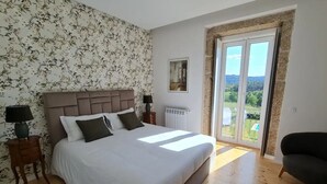 Ein Schlafzimmer mit einem großen Bett, einem Nachttisch mit Lampe, einem Sessel und Blick ins Freie durch eine Glastür.