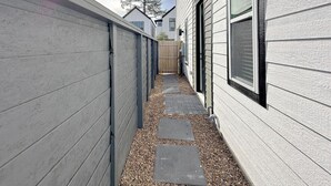 Un étroit passage entre deux bâtiments aux bardages gris, avec une clôture en bois et un chemin de gravier.