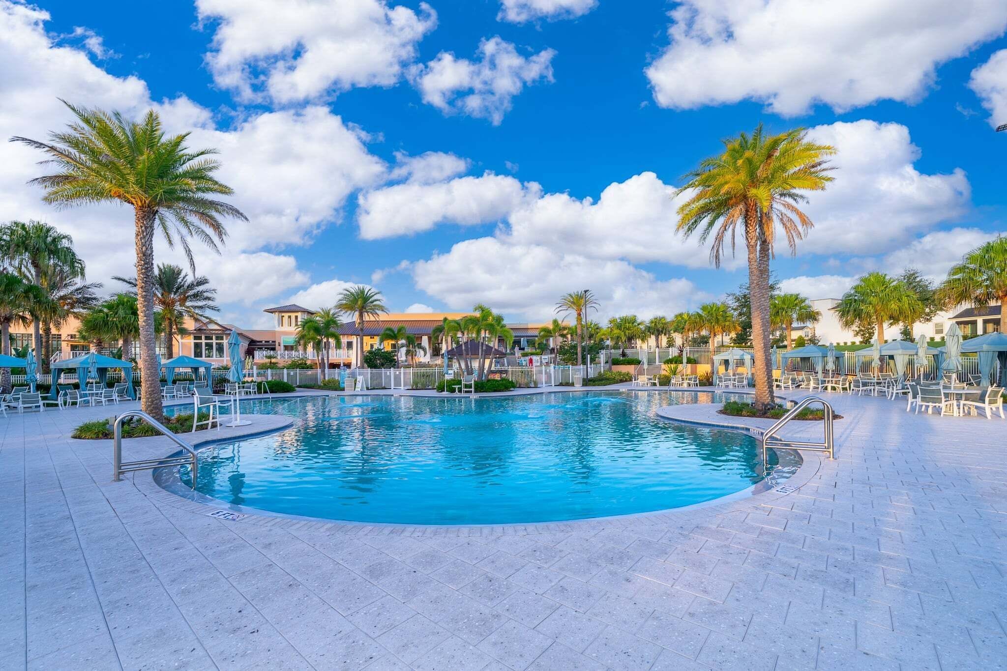 A swimming pool with lounge chairs and umbrellas, surrounded by palm trees and buildings.