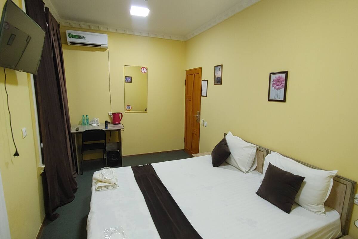 A hotel room with a bed, a desk, a chair, an air conditioning unit, a mirror, and a door.