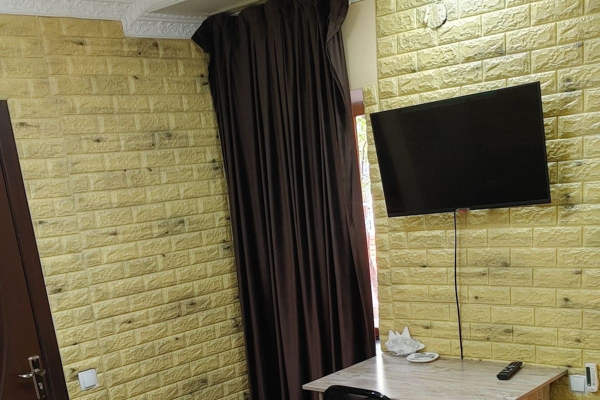 A hotel room with a brick-patterned wall, a flat-screen TV mounted on the wall, a wooden desk with a chair, and a bed with neatly folded towels.
