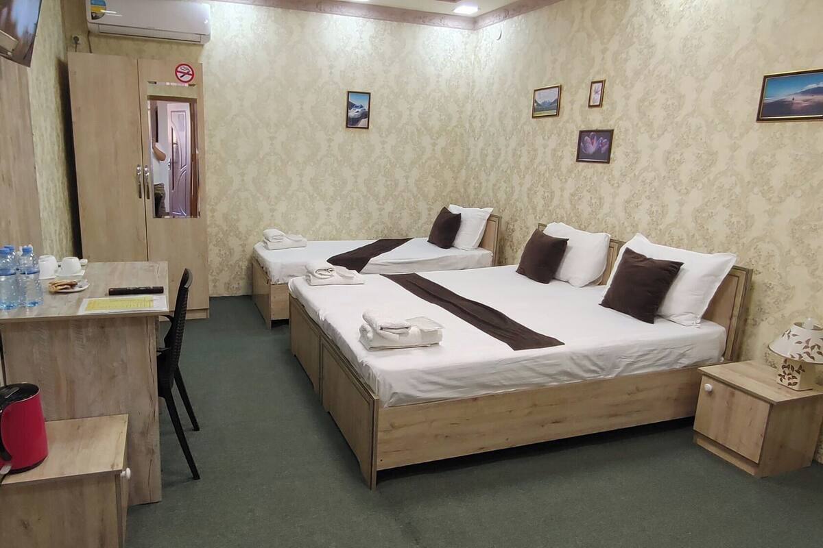 A hotel room with two beds, a desk with a chair, a radiator, and a small table with a lamp.