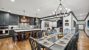 A modern kitchen with dark cabinets, a central island, and a dining area with a long table set for six. - North Nashville 15mins to Broadway Luxury Listing (Goodlettsville)