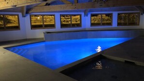 A swimming pool with blue lighting, wooden beams, and windows with snowy scenes.
