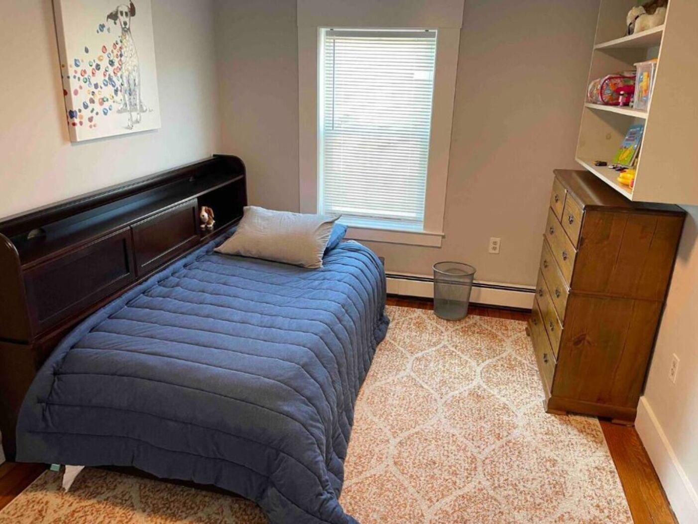 A bedroom with a bed, a window, a dresser, and a shelf with items.