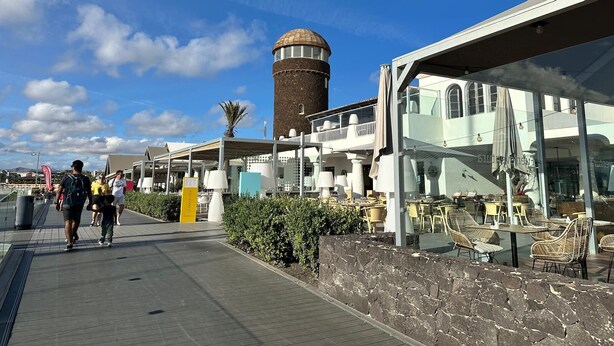 A waterfront promenade with a stone wall, outdoor seating, and a historic tower in the background. - Luxury 3 bedroom villa overlooking golf course with private pool and Wi-fi (Caleta De Fuste)