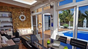 A modern living room with a stone wall, a glass pool, and a view of palm trees. - Luxury 3 bedroom villa overlooking golf course with private pool and Wi-fi (Caleta De Fuste)