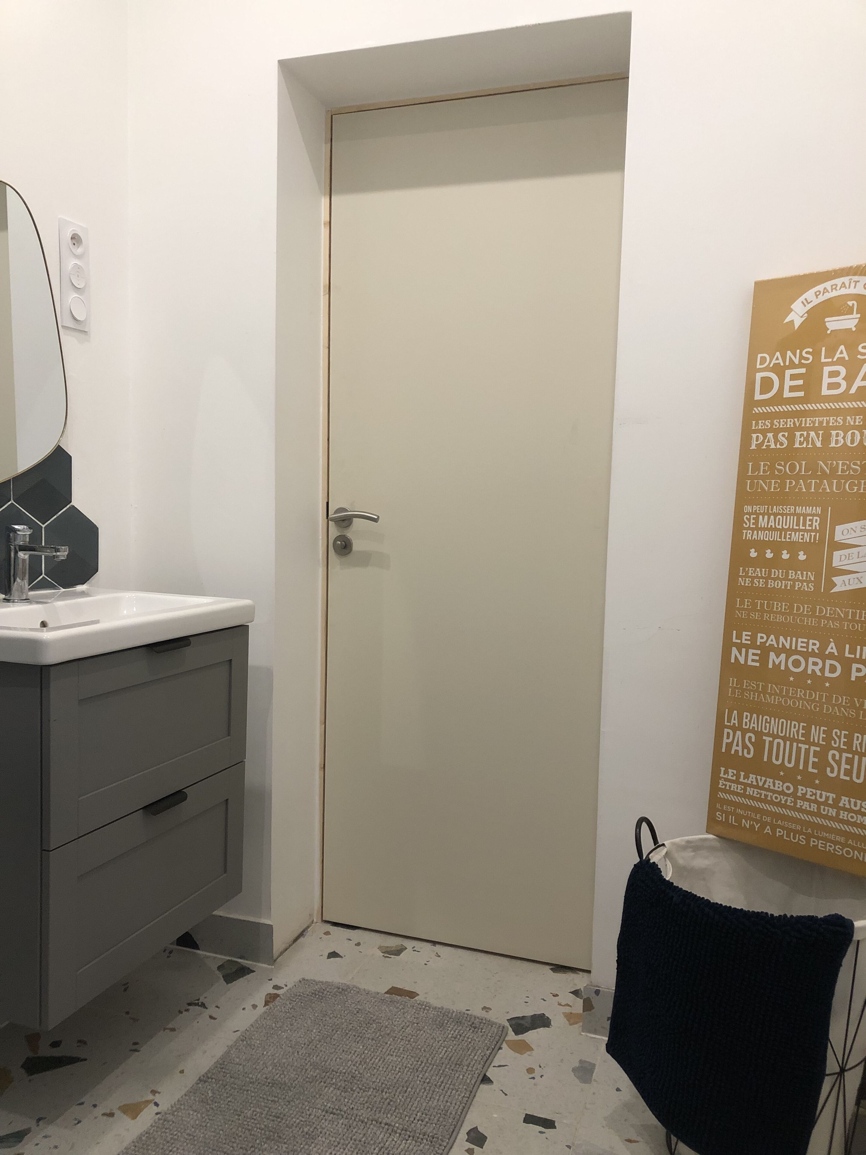 Une salle de bain avec un lavabo, un miroir et une porte. Il y a une affiche accrochée au mur et une serviette sur le sol.