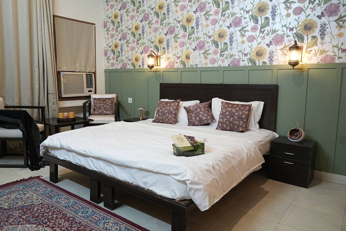 A neatly made bed with white linens, a floral wallpaper, and a green headboard. There are two bedside tables with lamps, a chair, and a small table with a yellow mug.