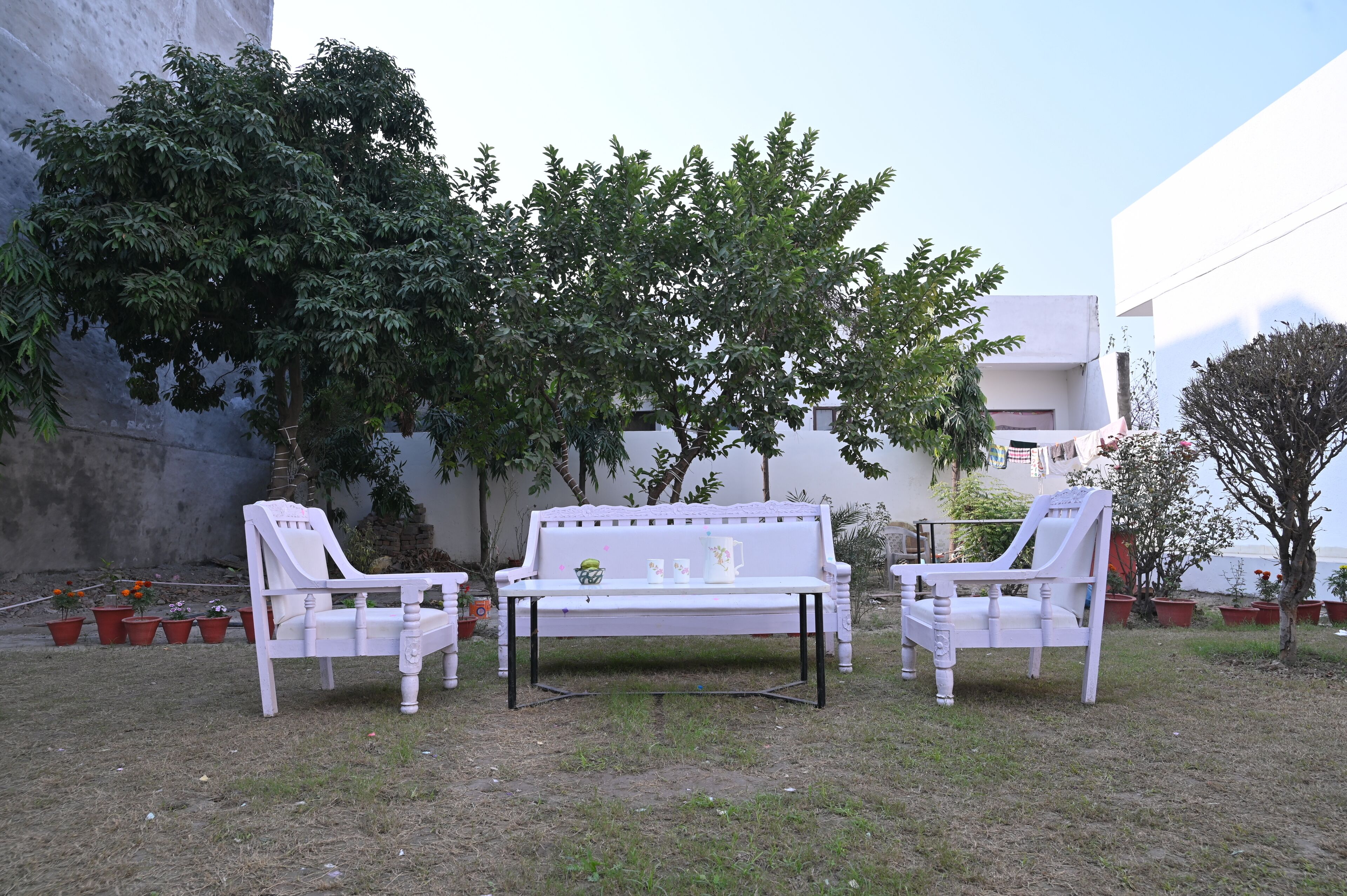 A white outdoor seating area with a bench and two chairs, surrounded by potted plants and trees.
