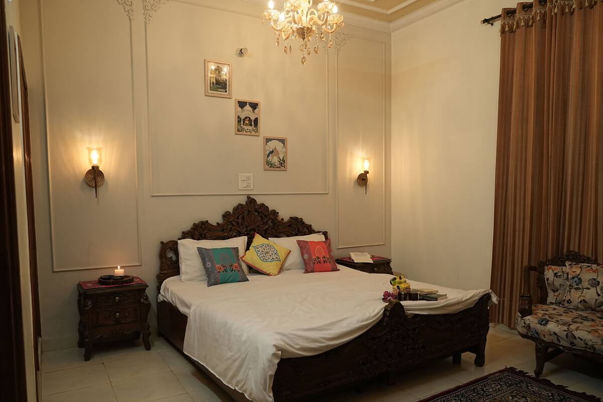 A bedroom with a large wooden bed, two bedside tables, a chandelier, wall-mounted lights, and a floral armchair.