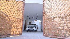 Ein weißer SUV ist vor einem Eingangstor mit aufwendiger Metallverzierung geparkt, im Hintergrund ist ein weißes Gebäude zu sehen.