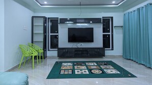 Een woonkamer met een flatscreen televisie, groene fauteuils, een kamerkleed met een patroon en een plafondventilator.