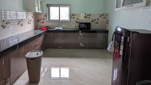 Een keuken met een raam, een magnetron, kastjes, een prullenbak en een koelkast.