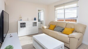 Ein Wohnzimmer mit beiger Couch, weißem Couchtisch und Fernseher auf einem Schrank. Auf dem Couchtisch steht eine kleine Pflanze, und es gibt eine Küchenzeile mit weißen Schränken und einem Fenster mit Jalousien.