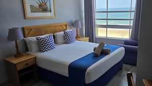 Habitación de hotel con cama, mesita de noche, lámpara y ventana con vista a la playa.