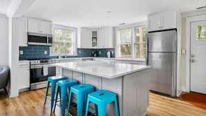 A modern kitchen with white cabinets, a central island with bar stools, a stainless steel refrigerator, and a large window providing natural light. - Folly Beach Compound - Near the Beach & Center St! (Folly Beach)