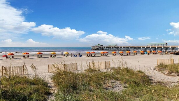 A beach with rows of umbrellas and chairs, a wooden pier with a gazebo, and a clear blue sky.