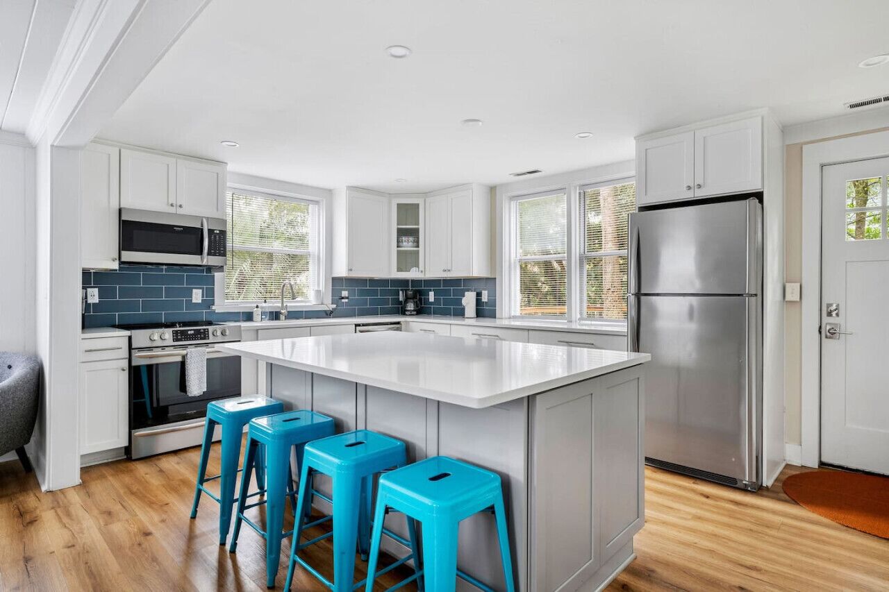A modern kitchen with white cabinets, a central island with bar stools, a stainless steel refrigerator, and a large window providing natural light.