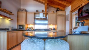 A modern kitchen with wooden cabinets, a stainless steel range hood, and a central island with two white furry stools. - Nicande - EXCEPTIONAL & LUXE chalet 8 pers (Haute-Nendaz)