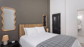 A modern bedroom with a wooden headboard, a large bed with white bedding, a plaid throw blanket, a nightstand with a lamp, and a mirror with a wavy frame.