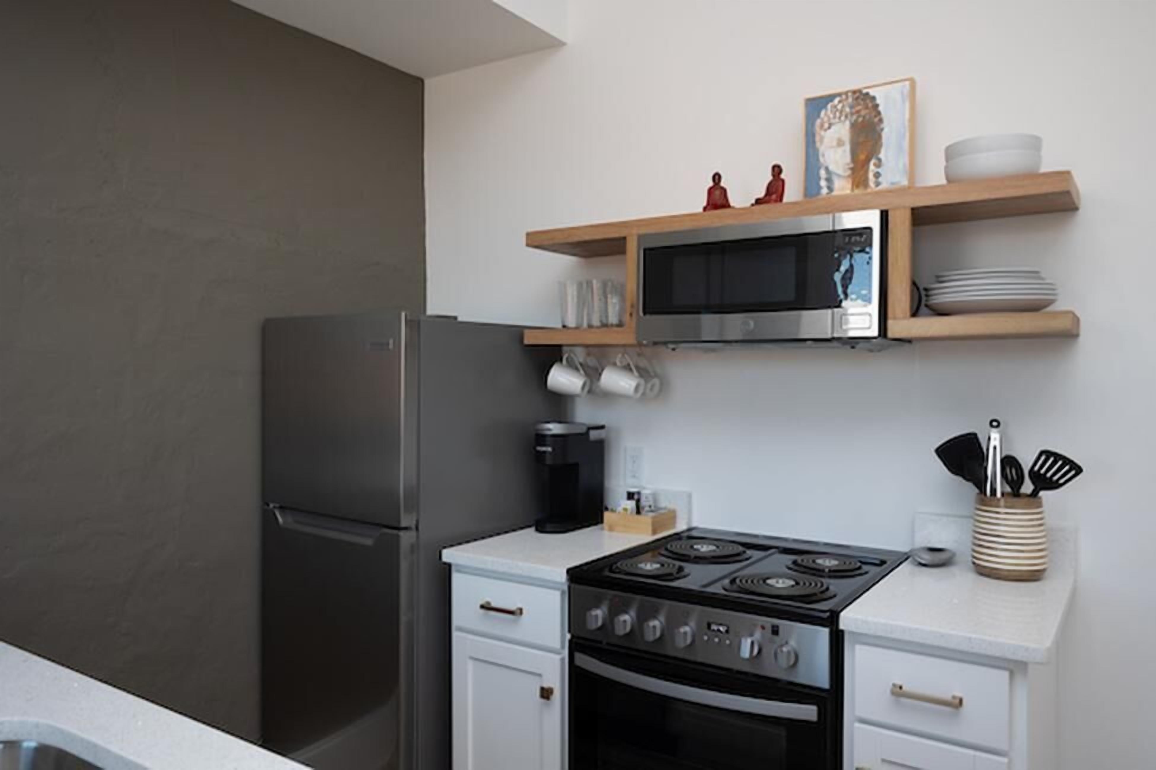 A compact kitchen with a stainless steel refrigerator, a microwave, a stove, and white cabinetry. There are wooden shelves above the stove holding dishes and a Buddha statue.