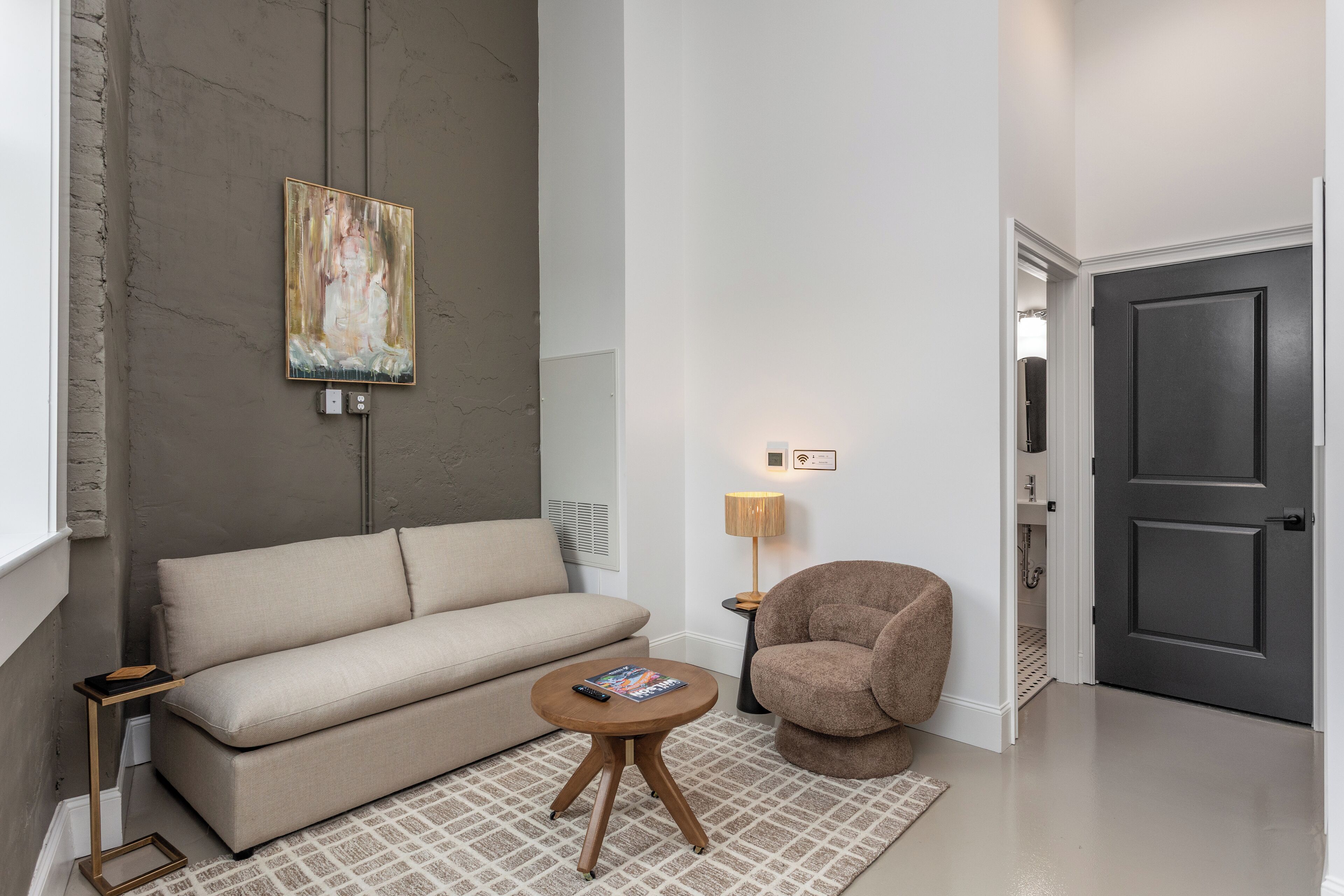 A modern living room with a beige sofa, a round wooden coffee table, a brown armchair, a painting on the wall, and a door in the background.