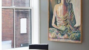 A room with a round wooden table, a large Buddha painting on the wall, and a view of a brick building through a window. - The Harriss (Wilson)