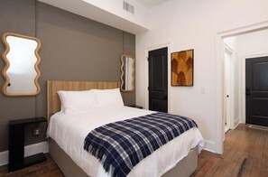 A bedroom with a wooden headboard, a white bed, a plaid blanket, two wall mirrors, and a door with a painting. - The Harriss (Wilson)