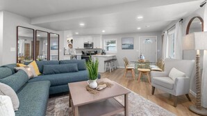 Ein modernes Wohnzimmer mit Ecksofa, Couchtisch und Essbereich mit Glastisch und Stühlen. Im Hintergrund ist eine Küche mit weißen Schränken und einer Mikrowelle erkennbar.