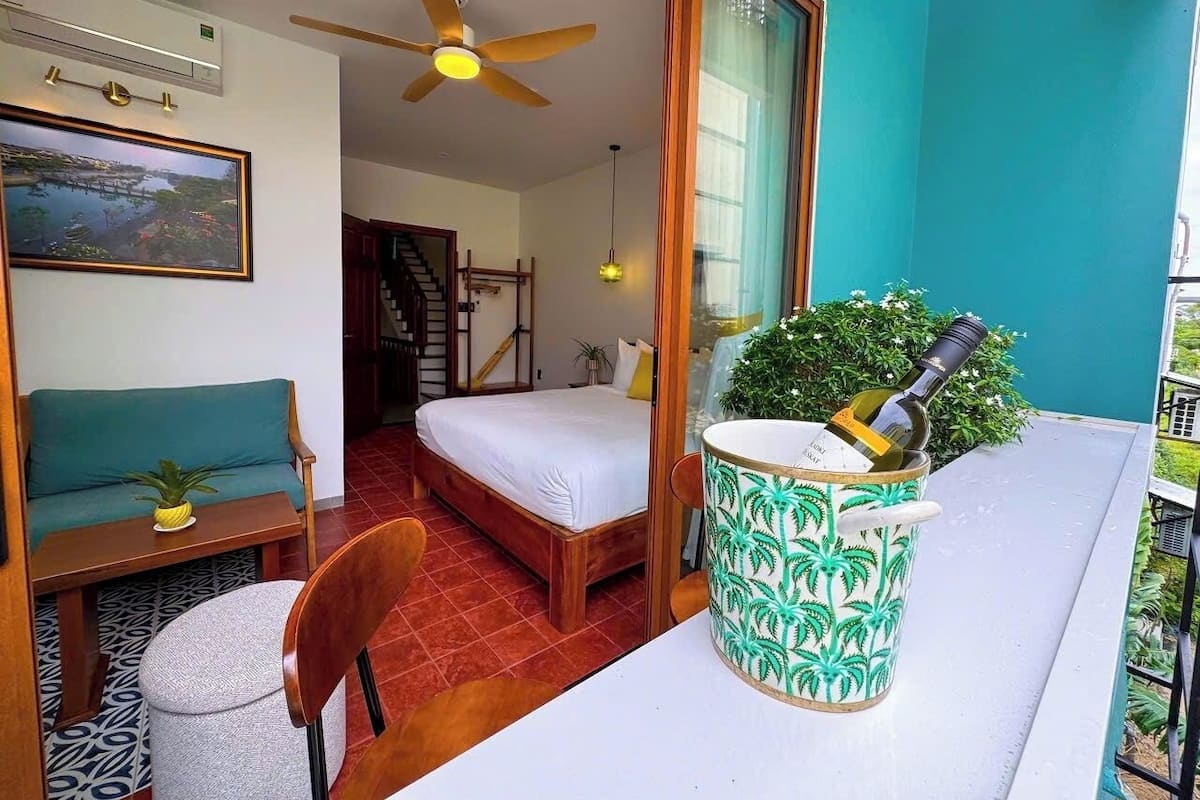 A room with a bed, a ceiling fan, a window, a small table with a plant, and a potted plant with a bottle on the balcony.