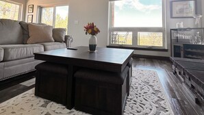 Ein geräumiges Wohnzimmer mit einer großen Couch, einem Couchtisch und einem Kamin. Mehrere Fenster sorgen für natürliches Licht und Blick ins Freie.