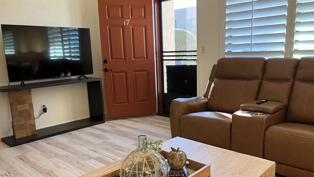 Un salón con sofá de cuero marrón, un televisor sobre un mueble negro, una mesa de centro de madera con objetos decorativos y una puerta con el número 17.