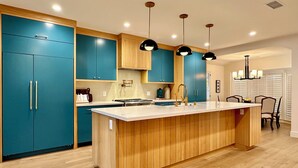 Une cuisine moderne avec des armoires de couleur turquoise, un îlot central en bois, des luminaires suspendus et un coin repas avec des chaises.