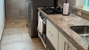 Cozinha moderna com geladeira de aço inoxidável, fogão a gás e forno embutido. Há uma janela com persianas, uma pia e bancada com porta-papel.