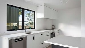 A modern kitchen with white cabinetry, a built-in dishwasher, and a window overlooking a backyard. - Modern Bach (Waihi Beach)