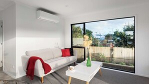 A modern living room with a white sofa, a red throw blanket, a white coffee table, and a large window offering a view of a backyard. - Modern Bach (Waihi Beach)