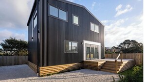 A modern two-story house with a dark exterior, large windows, and a wooden deck with steps leading to a glass door. - Modern Bach (Waihi Beach)